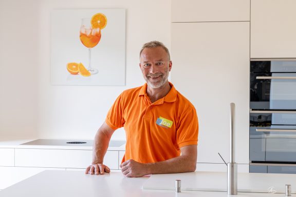 Ein Mann in orangefarbenem Shirt steht in einer modernen Küche.